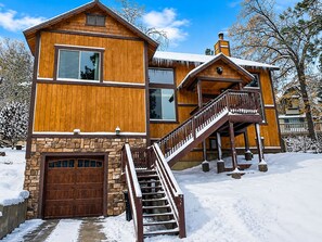 A two-story wooden house with a stone foundation, a wooden garage, and a staircase leading to a balcony. - Bear State of Mind - Close to Ski Slopes, Hot Tub, Foosball, Fireplace, Deck, BBQ (Big Bear City)