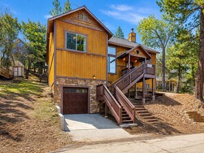 A two-story wooden house with a stone foundation, a front garage, and a staircase leading to a second-floor balcony. - Bear State of Mind - Close to Ski Slopes, Hot Tub, Foosball, Fireplace, Deck, BBQ (Big Bear City)