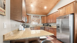 A kitchen with wooden cabinets, a stainless steel refrigerator, granite countertops, and bar stools. - Bear State of Mind - Close to Ski Slopes, Hot Tub, Foosball, Fireplace, Deck, BBQ (Big Bear City)