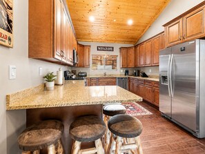 A kitchen with wooden cabinets, a stainless steel refrigerator, granite countertops, and a bar stool area. - Bear State of Mind - Close to Ski Slopes, Hot Tub, Foosball, Fireplace, Deck, BBQ (Big Bear City)