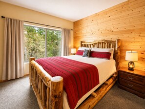 A wooden-framed bed with a red and white bedspread, two bedside lamps, and a view of trees through a large window. - Bear State of Mind - Close to Ski Slopes, Hot Tub, Foosball, Fireplace, Deck, BBQ (Big Bear City)