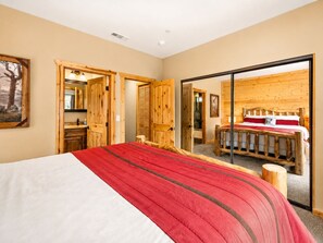 A bedroom with a large bed, wooden walls, and a sliding door leading to another room. - Bear State of Mind - Close to Ski Slopes, Hot Tub, Foosball, Fireplace, Deck, BBQ (Big Bear City)