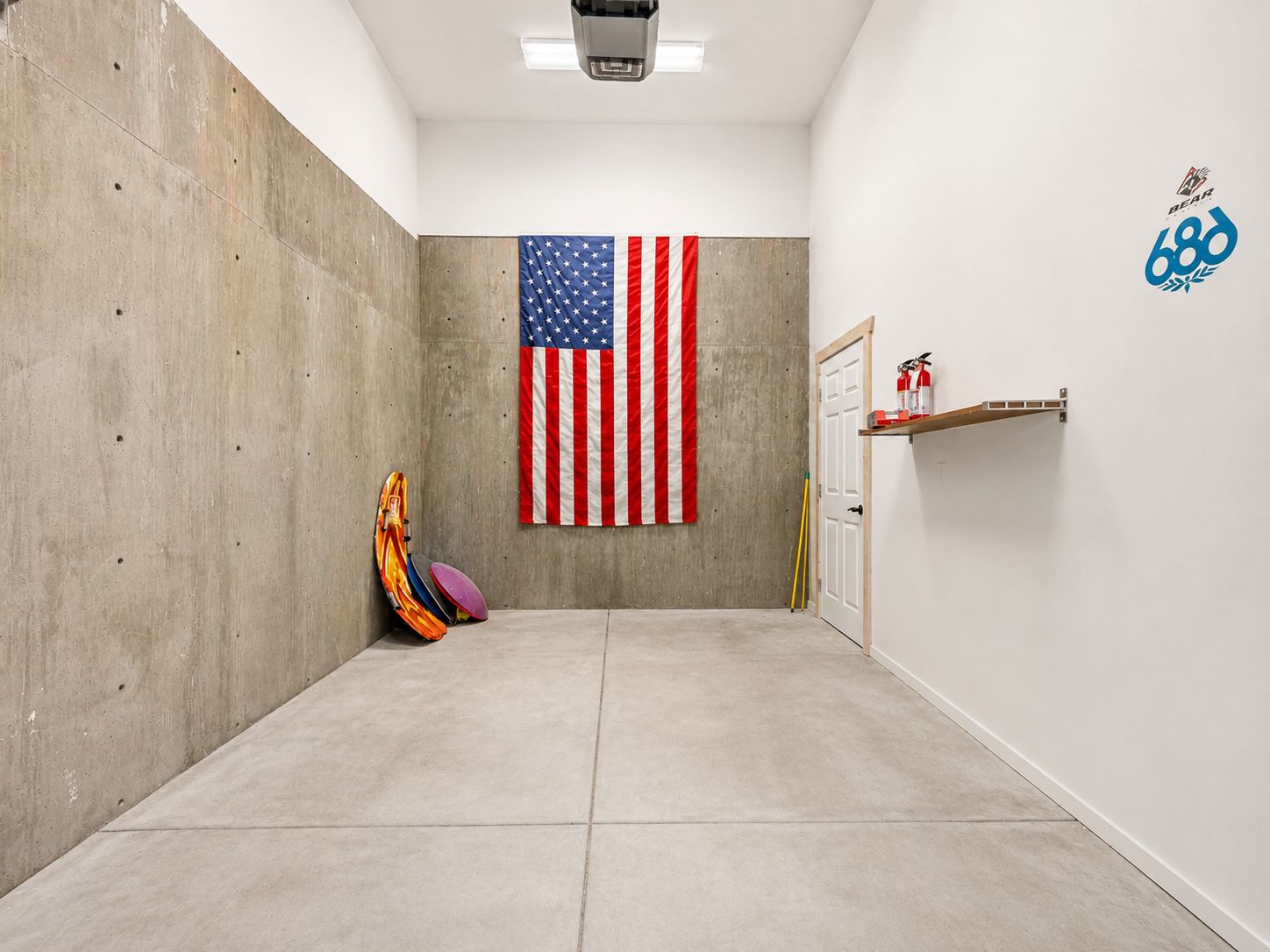 A hallway with a concrete wall, a mounted American flag, a door, and a shelf with fire extinguishers.