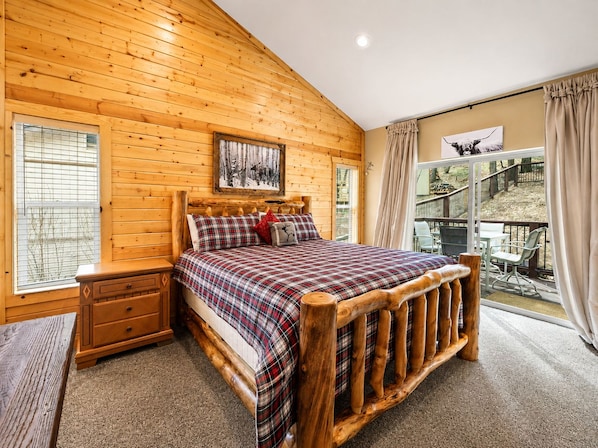 A wooden cabin bedroom with a bed, nightstand, and a view of a deck. - Bear State of Mind - Close to Ski Slopes, Hot Tub, Foosball, Fireplace, Deck, BBQ (Big Bear City)