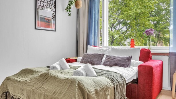 Una cama bien hecha con una manta verde, almohadas blancas y una silla roja. Un cuadro enmarco cuelga de la pared, y una planta está en el alféizar de la ventana.