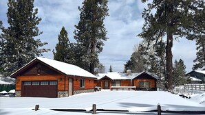 A wooden cabin with a garage, surrounded by snow-covered ground and tall pine trees. - Log Cabin Dream - Log Cabin, Walk to Lake, Fireplace, Deck, BBQ (Big Bear Lake)