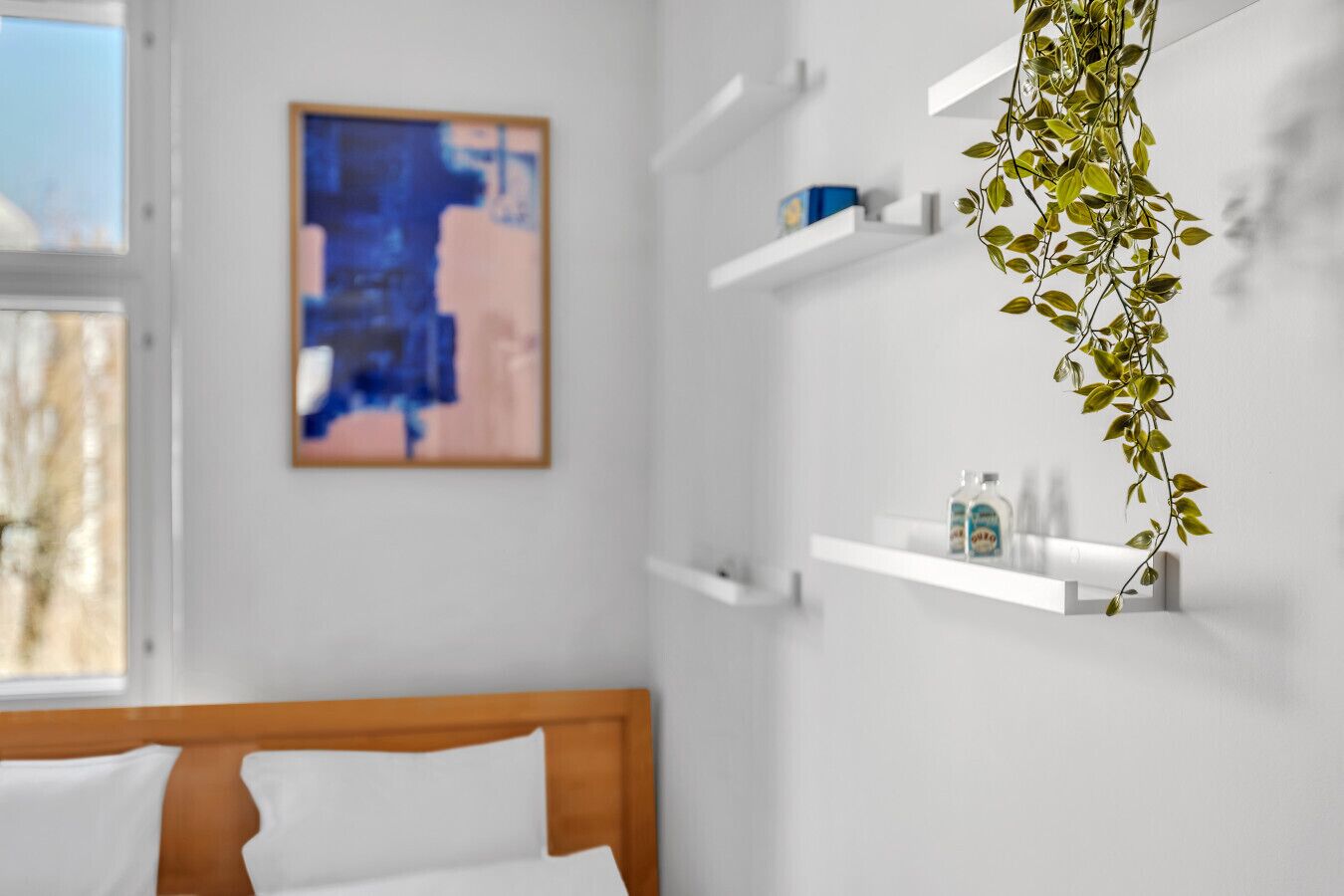 Une chambre avec une tête de lit en bois, une plante suspendue, un tableau au mur et des étagères blanches où sont rangés les articles de toilette.