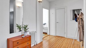 Een kamer met een houten vloer, witte muren, een houten dressoir met een plant, een radiator en een kapstok met een jas.