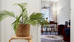 A room with a woven plant pot on a wooden stool, a dining area with chairs, a red sofa, and a patterned rug. - Sunny Views | Design District | Balcony | WiFi (Helsinki)