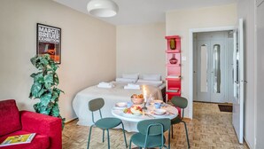 A compact hotel room with a bed, a dining table with chairs, a red armchair, a potted plant, and a poster on the wall. - Vibrant & Central w Netflix, WiFi, 200m tram/bus (Helsinki)