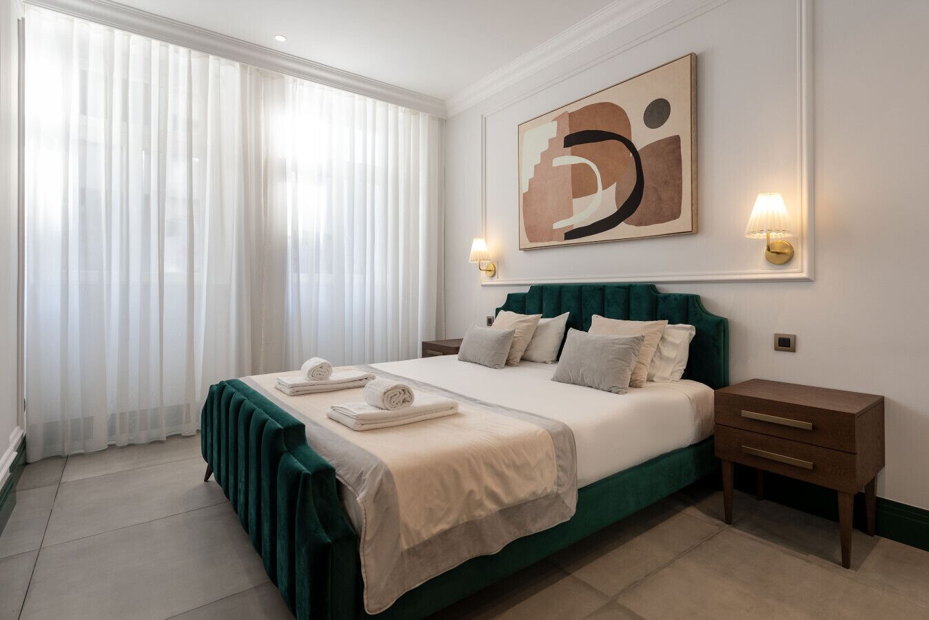 A hotel room with a large bed, two bedside tables, a wall-mounted artwork, and a window with sheer curtains.
