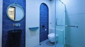 Et badeværelse med blå fliservæg, et rundt spejl, en sort vask, et toilet og en brusekabine i glas.