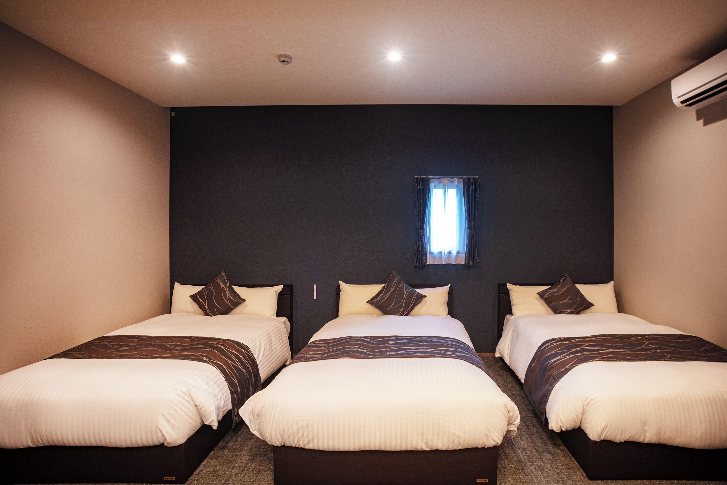 A hotel room with three beds, a dark wall, a window with curtains, and an air conditioning unit.