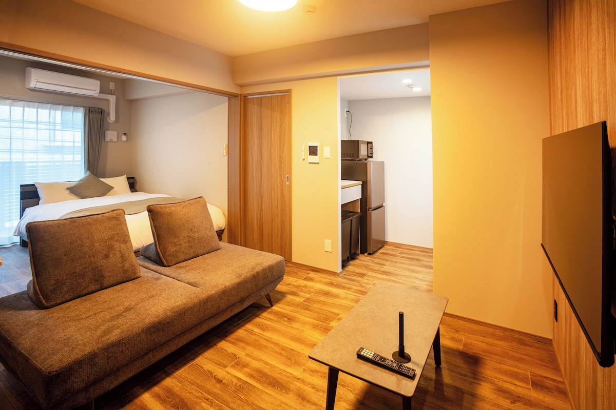 Una habitación de hotel compacta con sofá, una mesita con un control remoto, televisor, cama y kitchenette.