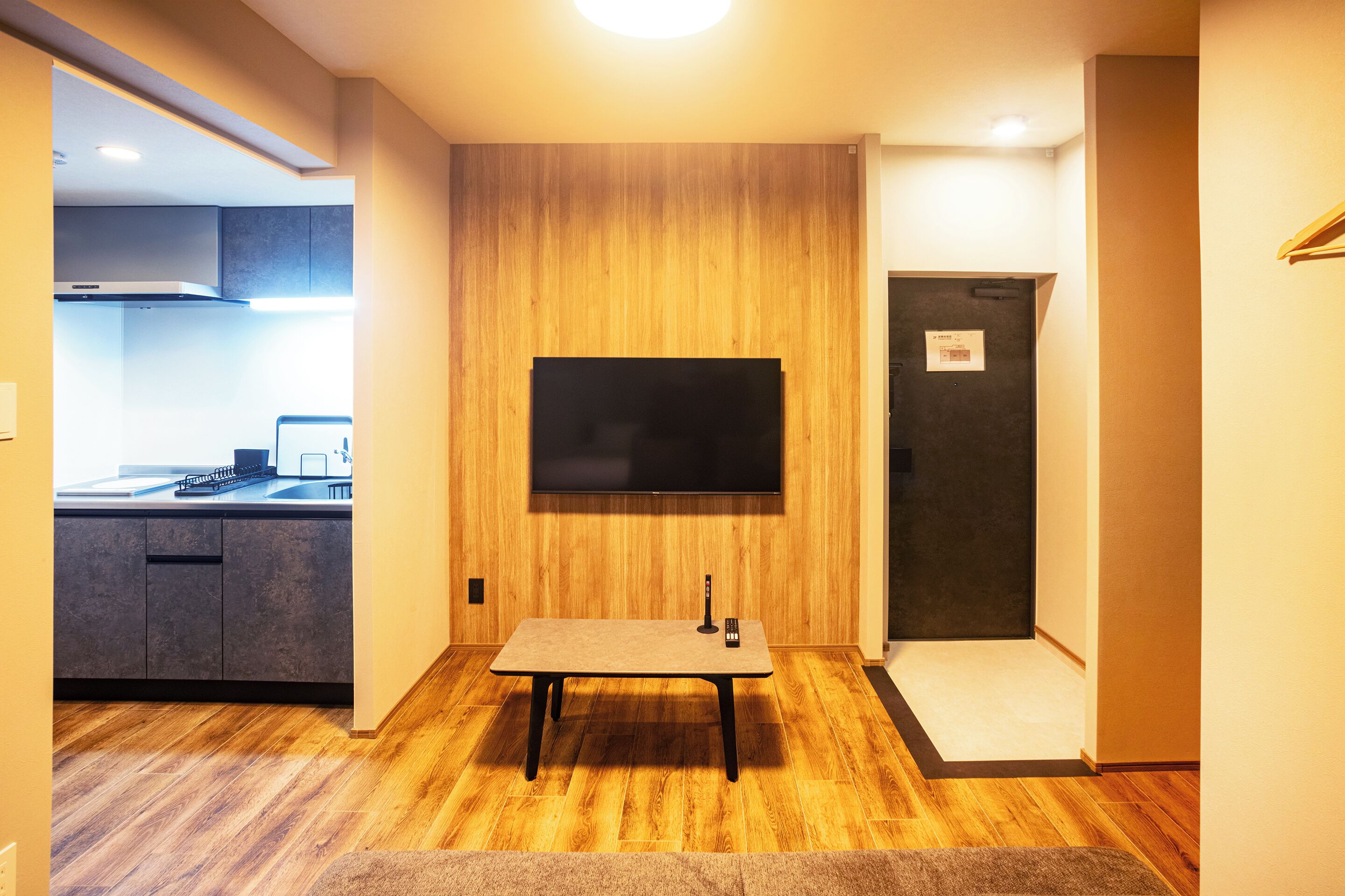 A modern room with a wooden panel wall, a flat-screen TV mounted above a small table, and a kitchen visible through an open door.