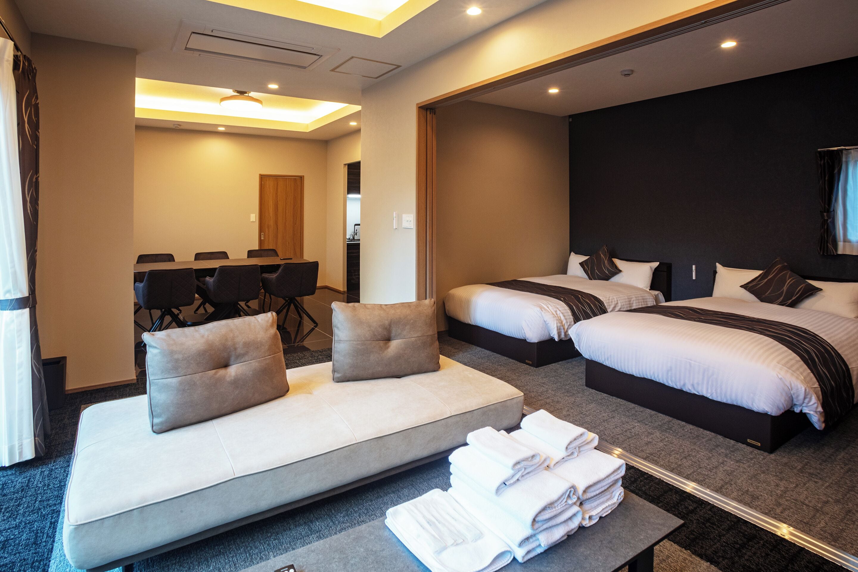 A hotel room with a sofa, two beds, a dining table, and a bathroom.