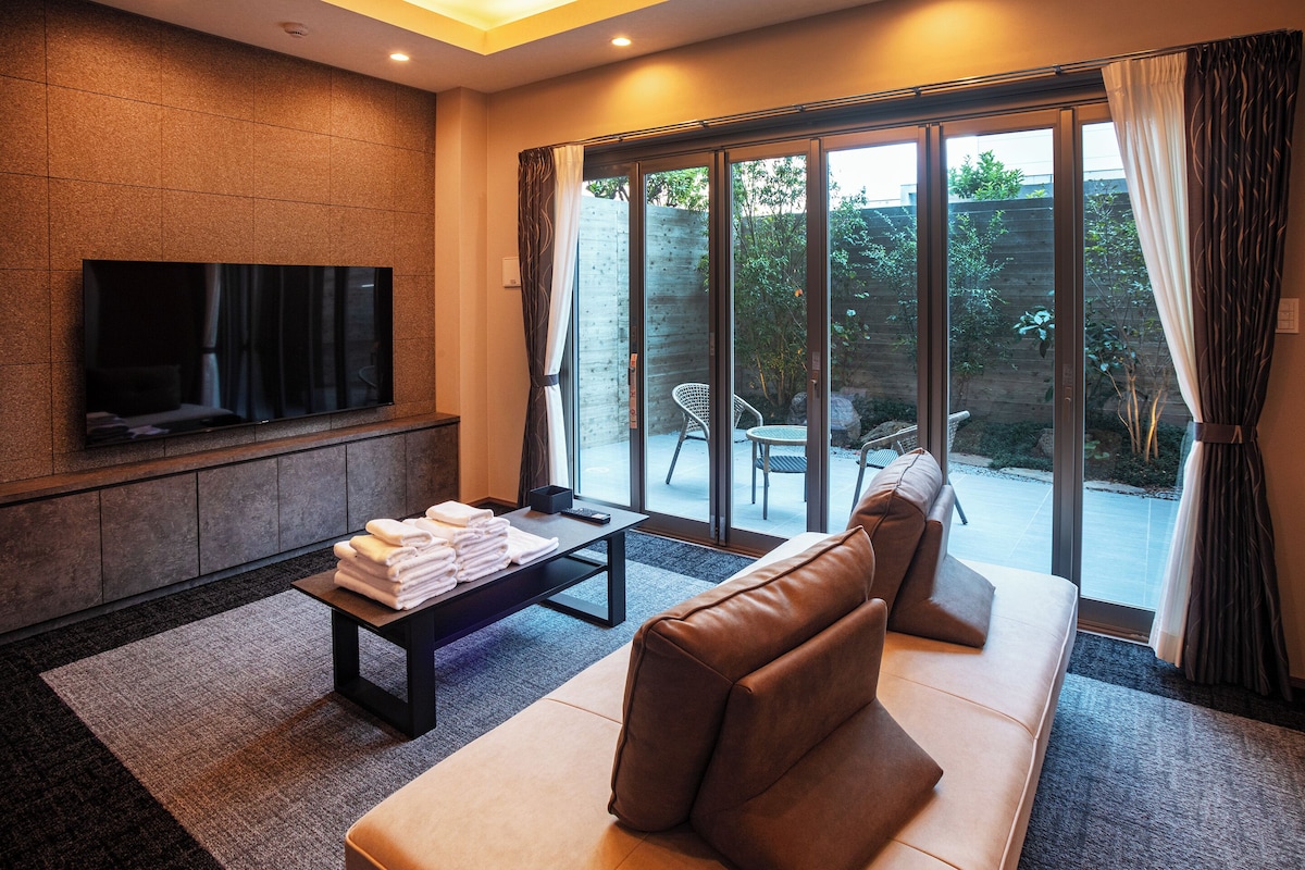 Una sala de estar moderna con una televisión de pantalla plana, un sofá, una mesa de centro y vistas a un jardín a través de puertas correderas de vidrio.
