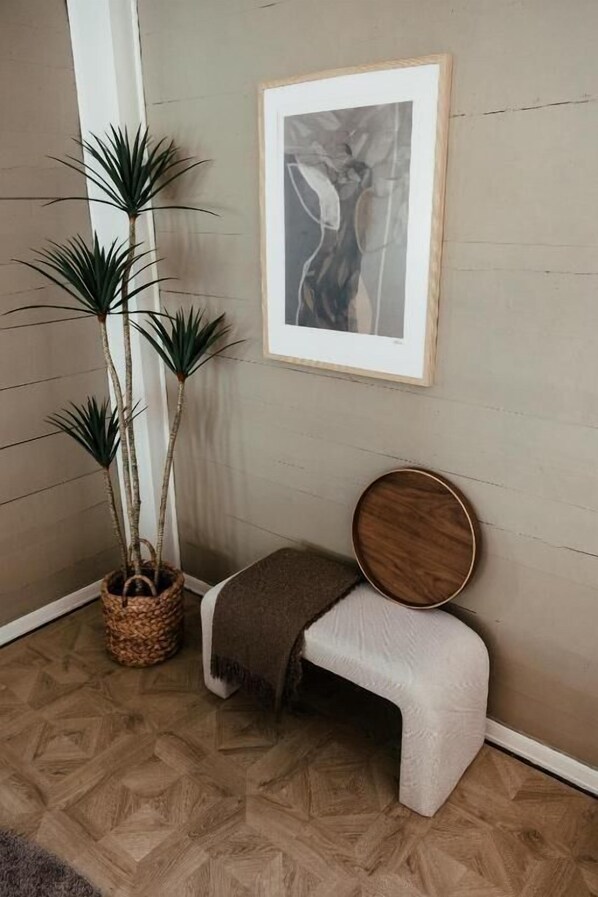 A corner room with a herringbone wooden floor, a white bench with a brown cushion, a framed picture on the wall, and two tall potted plants. - Huset Vårt Bed and Breakfast (Volda)