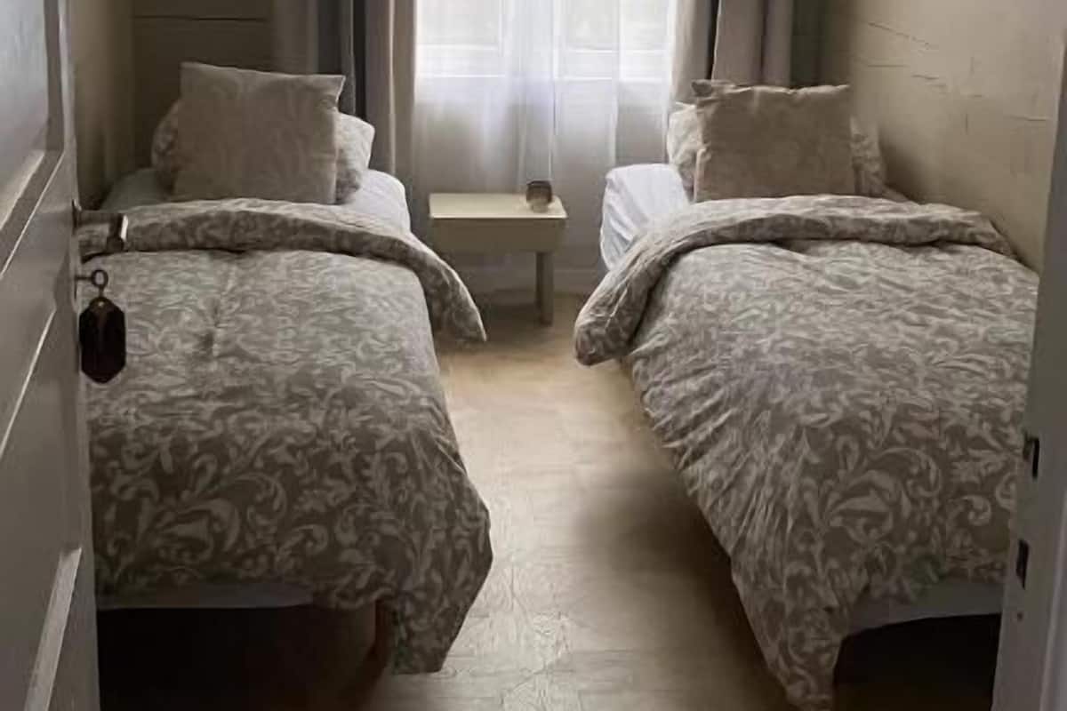 A small, narrow room with two single beds, a small table, and a window with sheer curtains.