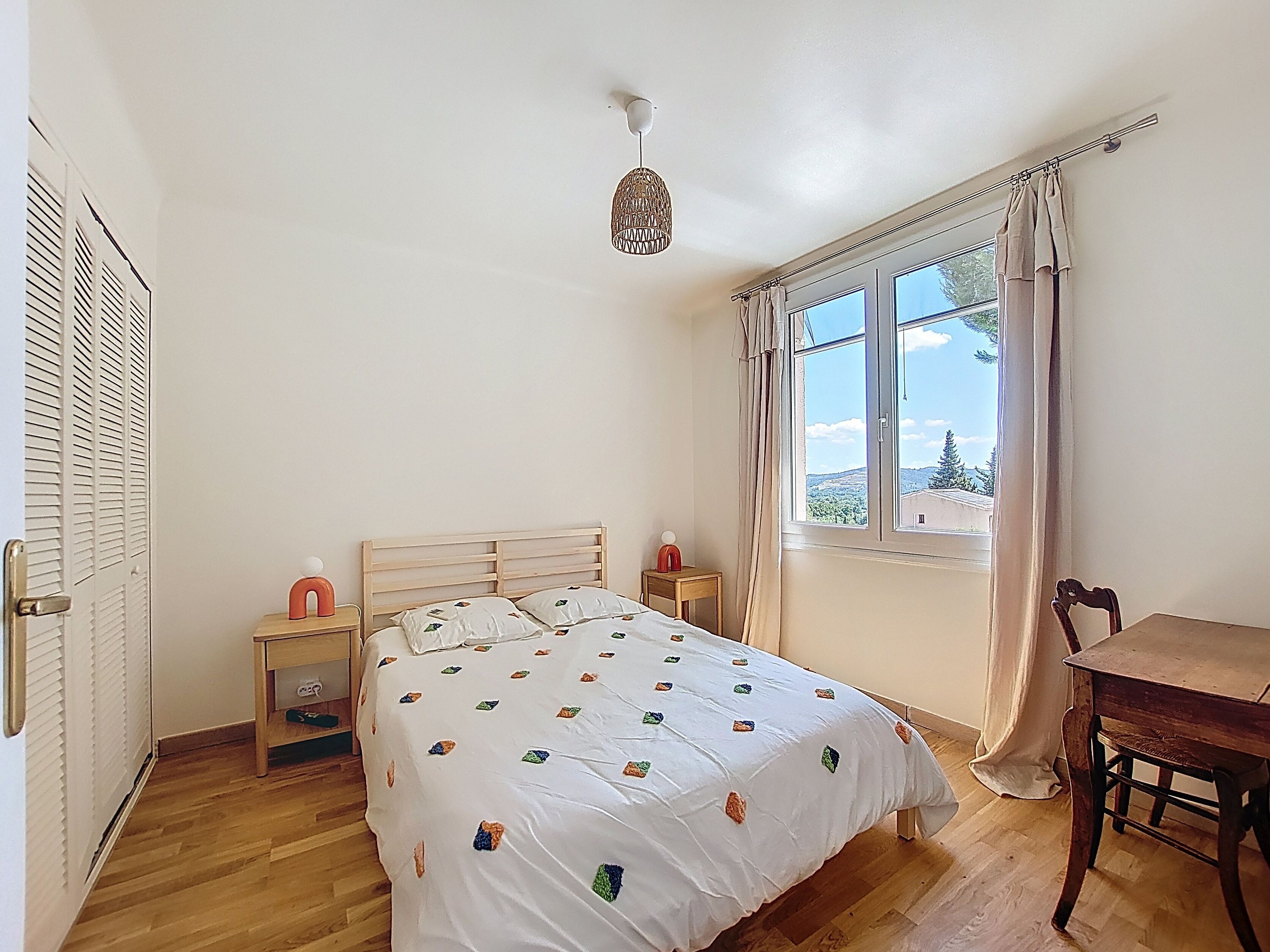 A bedroom with a wooden bed, a bedside table, a chair, a window with curtains, and a view of the outdoors.