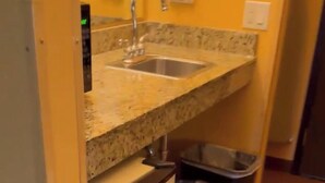 Une cuisine compacte dotée d’un plan de travail en granit, d’un évier en acier inoxydable et d’un petit réfrigérateur. On y trouve deux poubelles et un distributeur de savon fixé au mur.