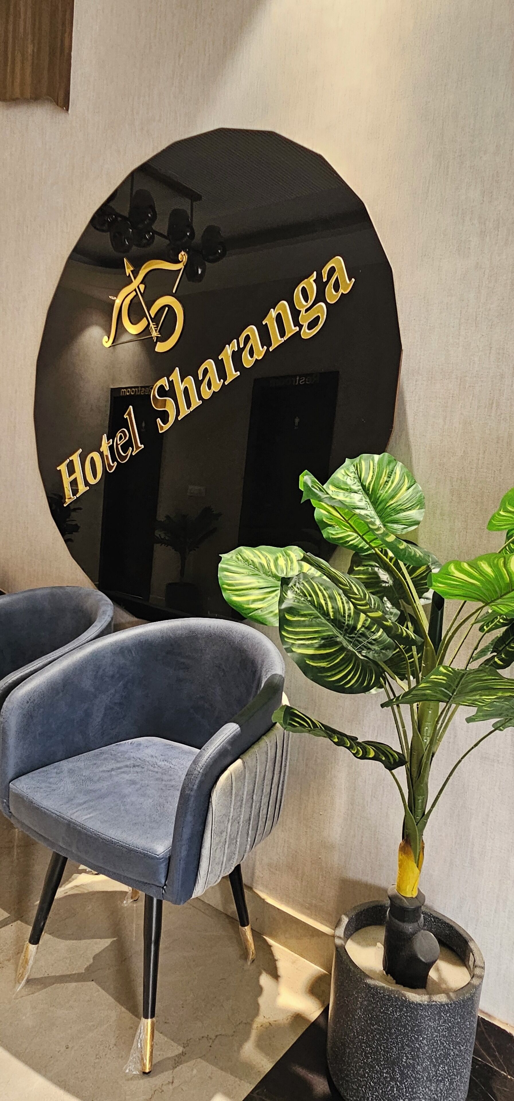 A modern hotel interior with a large black sign reading "Hotel Sharanga," a blue armchair, and a potted plant.