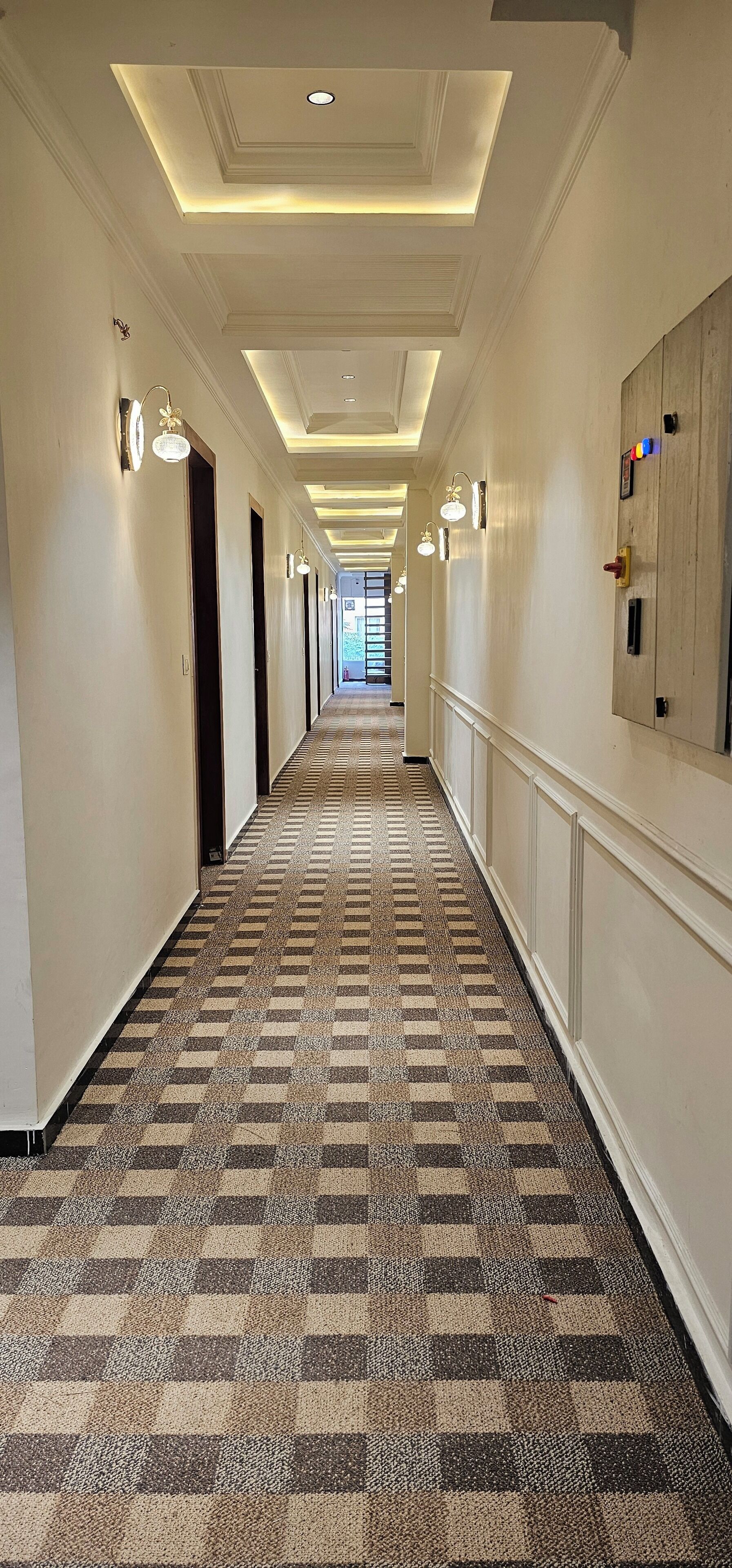 Un couloir d’hôtel avec un tapis à carreaux, un éclairage encastré et une porte en bois avec une serrure située à droite.