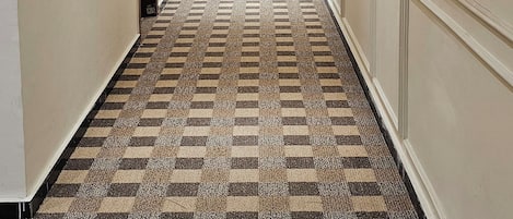 A hotel hallway with a checkered carpet, recessed lighting, and a wooden door with a lock on the right.