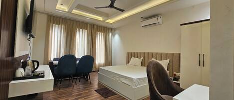 A hotel room with a bed, a desk with chairs, a television, an air conditioning unit, and a ceiling fan.