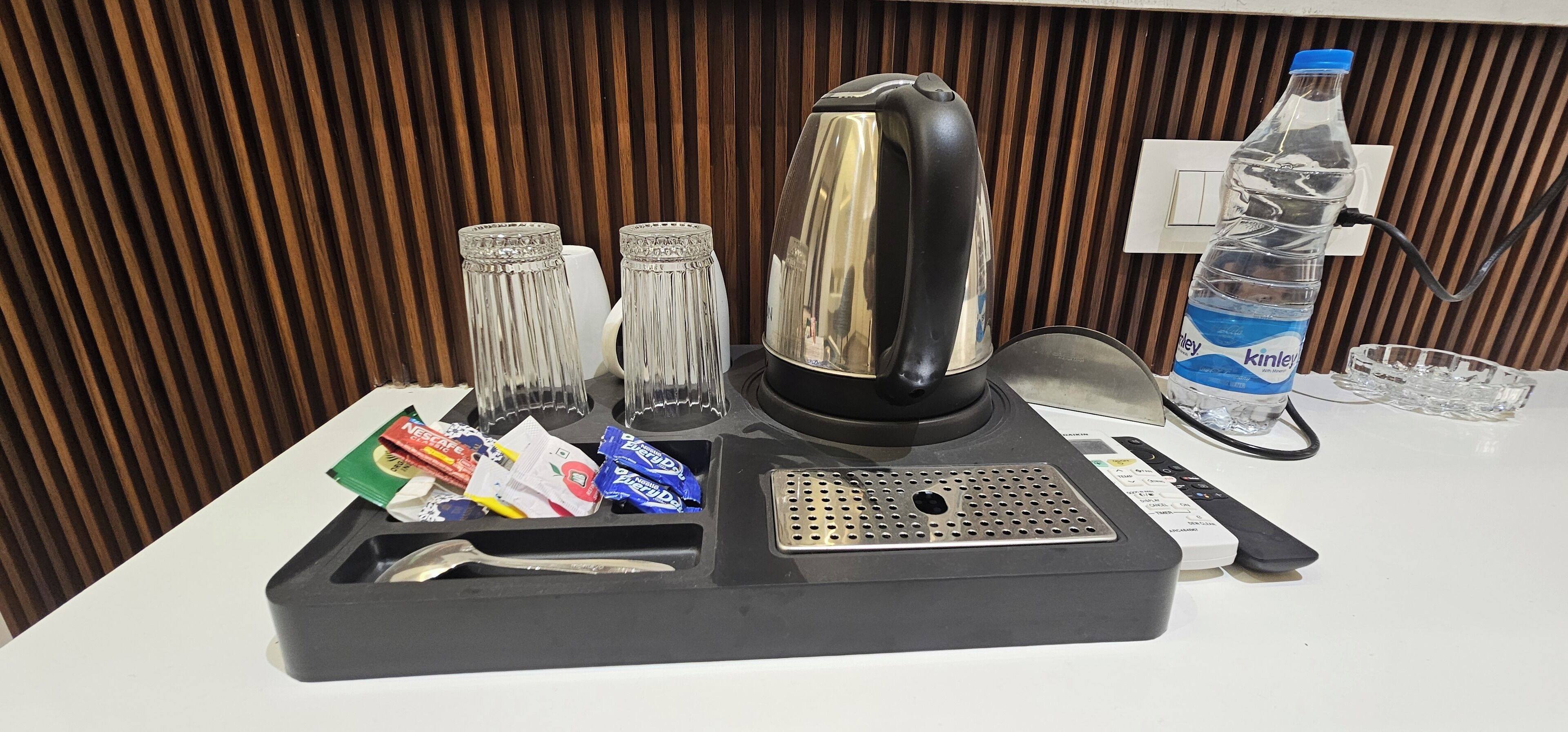 A hotel minibar setup with a kettle, glasses, a bottle of water, and various snacks.