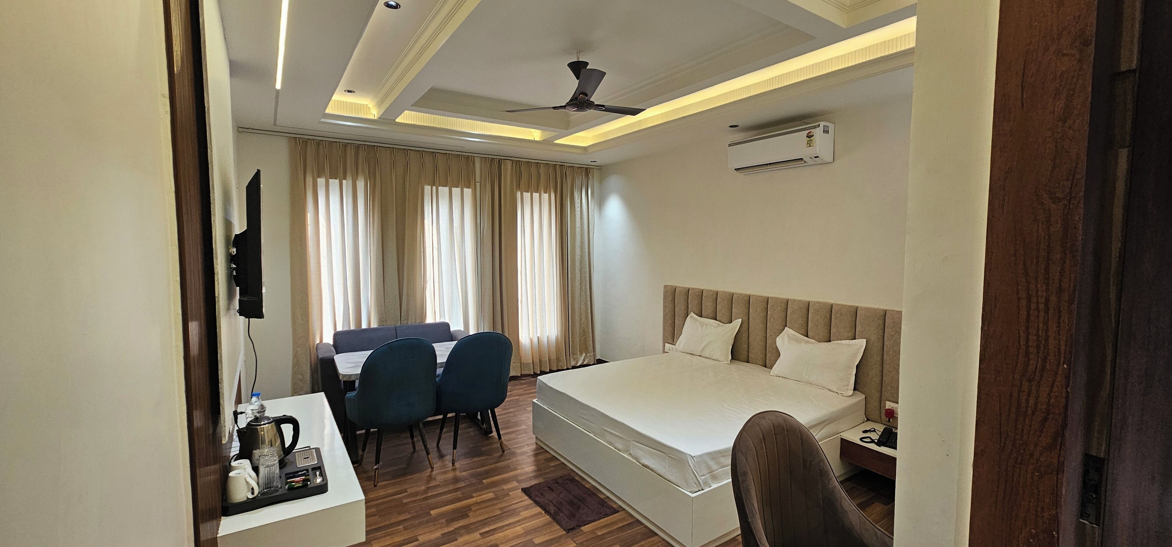 A hotel room with a bed, a desk with a chair, a dining table with chairs, and an air conditioning unit.