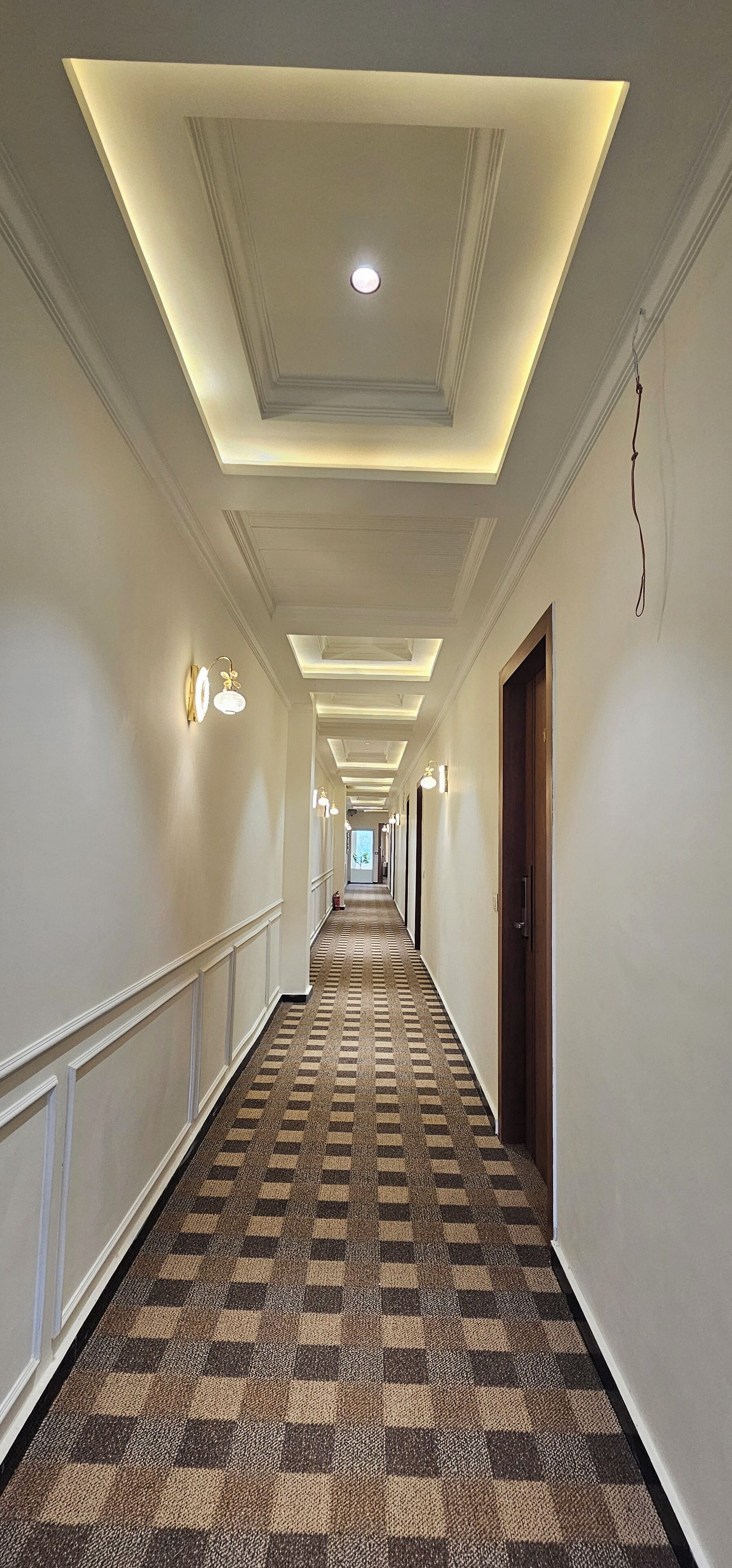 A hotel hallway with a checkered carpet, recessed lighting, and wall sconces.