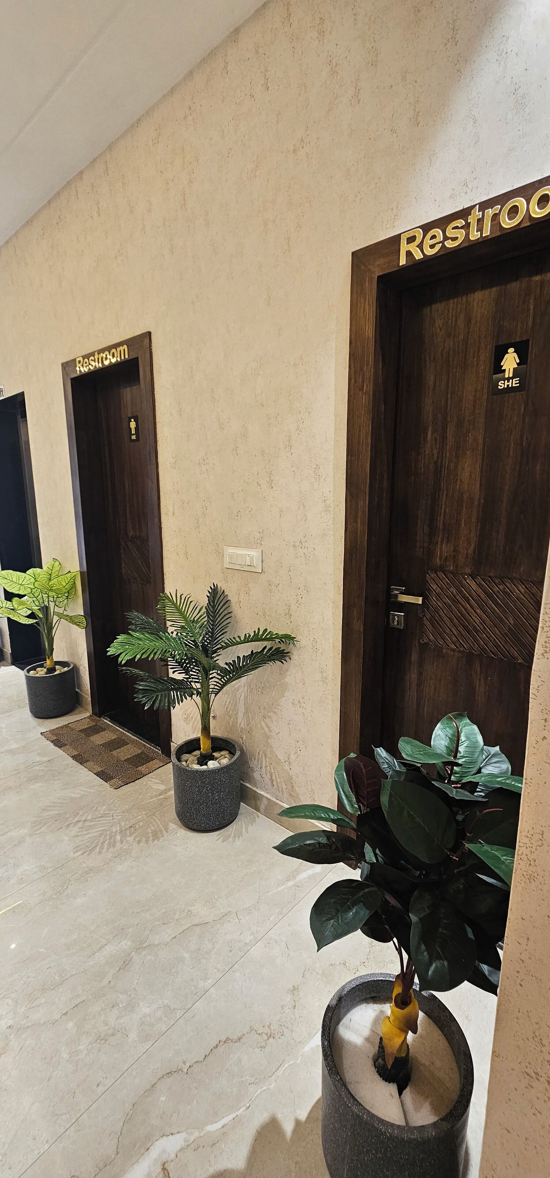 A hallway with two doors labeled "Restroom" and "SHE," each with a corresponding restroom symbol. There are potted plants along the hallway.