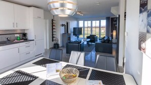 A modern kitchen with white cabinets, a dining area with a checkered tablecloth, and a living room with a TV and seating. - Reethaus Arkonablick - Villa by the sea with fenced garden. Dogs welcome. (Glowe)