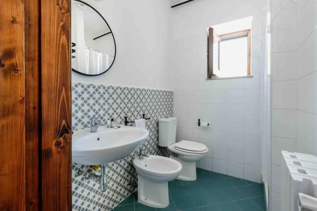 A bathroom with a white sink, a bidet, and a toilet. There is a window on the wall and a wooden door frame.