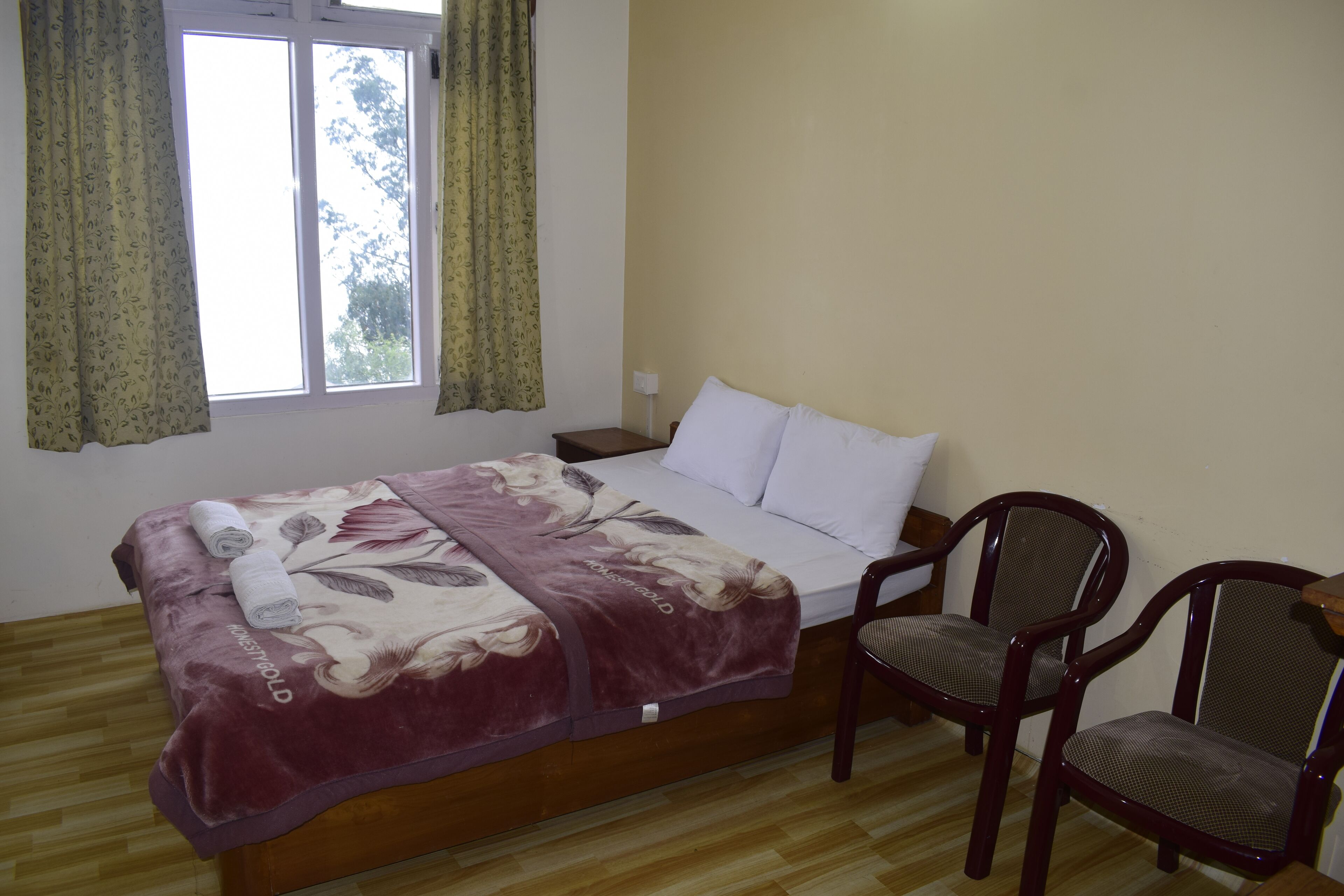 A bedroom with a bed, two chairs, a window with curtains, and a wooden floor.