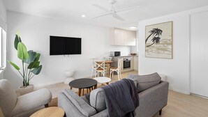 Ein modernes Wohnzimmer mit einem grauen Sofa, einem Couchtisch und einem Essbereich mit weißem Tisch und Stühlen. Ein Deckenventilator ist sichtbar, und in der Ecke steht eine große Pflanze.