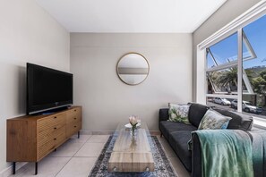 Un salon moderne avec un téléviseur à écran plat posé sur un meuble en bois, un grand miroir rond accroché au mur, un canapé avec des coussins et une grande fenêtre donnant sur la rue.