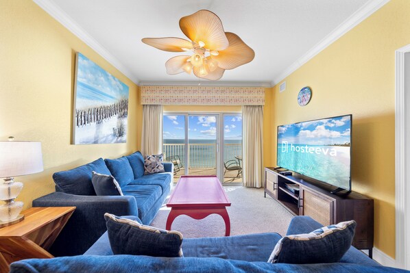 A living room with a sofa, a coffee table, a TV, and a balcony with a view. - Crystal Tower 1802 (Gulf Shores)