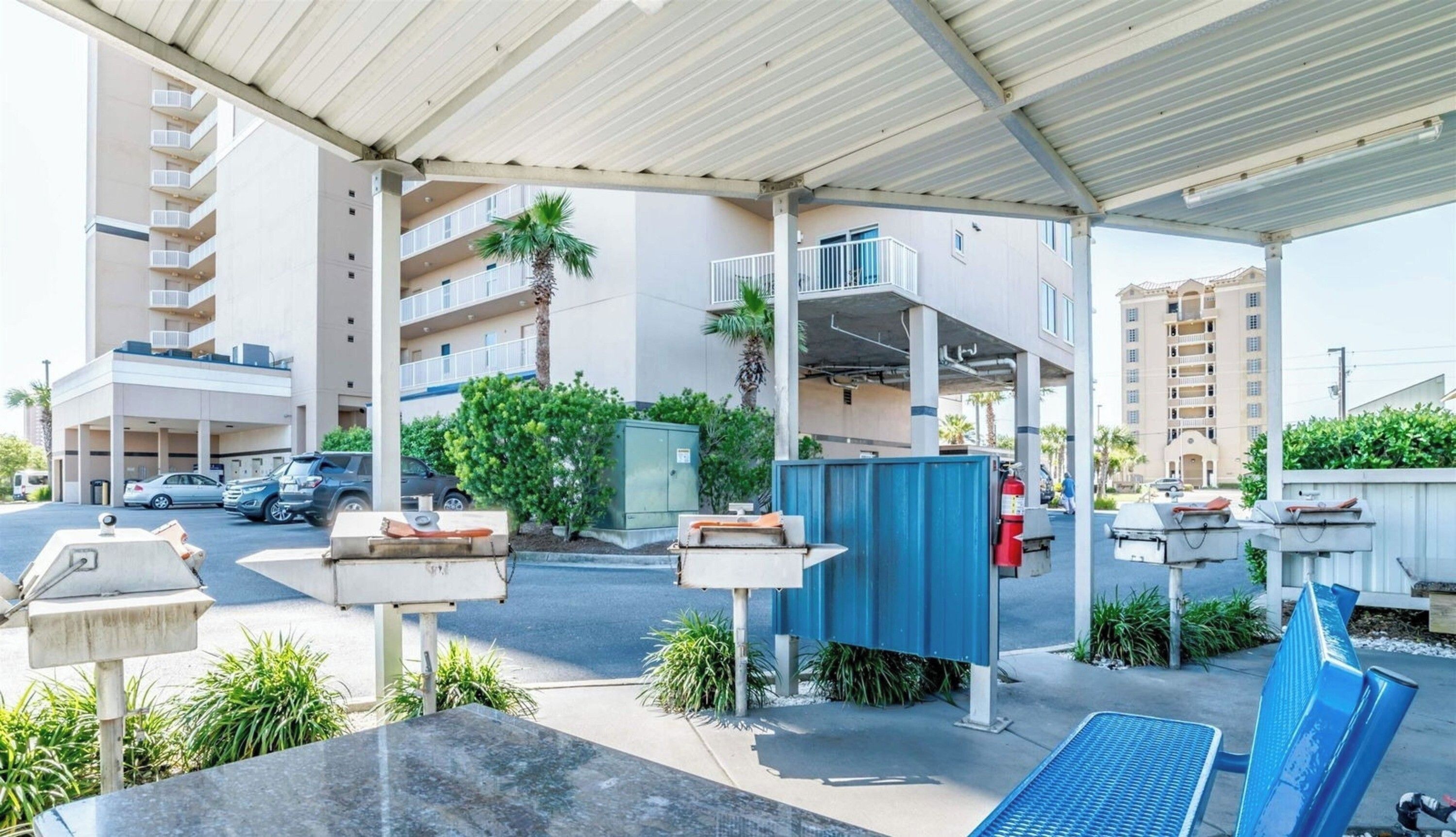 A covered parking area with a building in the background, a fire extinguisher, and a blue trash bin.