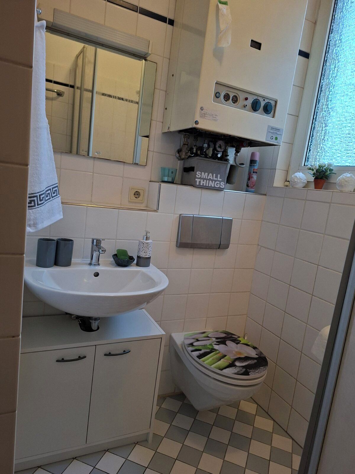 Un baño pequeño con lavamanos blanco, espejo e inodoro. Hay una toalla colgada en la puerta y un pequeño estante con artículos de tocador.