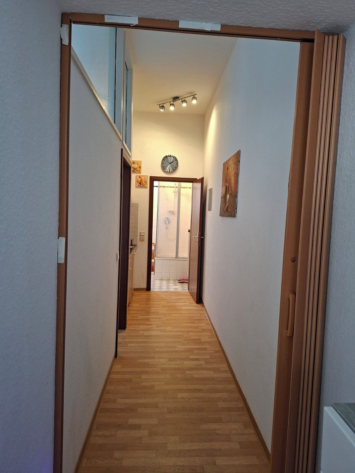 A narrow hallway with wooden flooring, a ceiling light fixture, and a clock on the wall.
