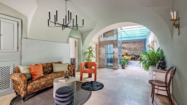 A spacious living area with a high ceiling, a chandelier, a sofa, a coffee table, and a view through an archway to a dining area. - Giselle Buda Castle (Budapest)