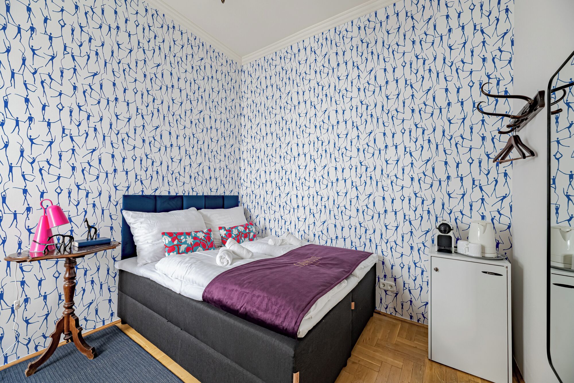 A bedroom with a patterned wallpaper, a bed with a blue headboard, a small table with a lamp, and a refrigerator.