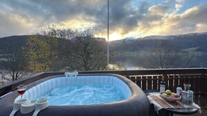 Una vasca idromassaggio con vista sulle montagne e un lago, immersa tra alberi e un pontile di legno.