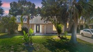 Une maison de plain-pied avec une façade en briques, une porte d’entrée et un petit jardin devant, agrémenté d’un palmier et d’une voiture garée dans l’allée.