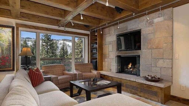 A cozy living room with a stone fireplace, a large window with a view of trees, and a comfortable seating area. - Treetops 4e 3 Bedroom Condo (Avon)