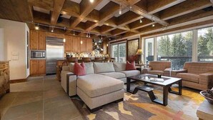A spacious living area with a large sectional sofa, a coffee table, and a fireplace. There is a kitchen area with wooden cabinets and a stainless steel refrigerator. The room has large windows offering a view of the outdoors. - Treetops 4e 3 Bedroom Condo (Avon)