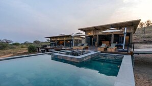 A pool with lounge chairs and umbrellas, a building with a sloped roof, and a view of the surrounding landscape.