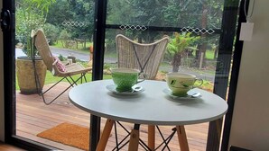 A round table with two green cups on saucers, a wicker chair, and a view of a garden through a glass door. - Watts River Tiny - Hidden, but Close to Township (Healesville)
